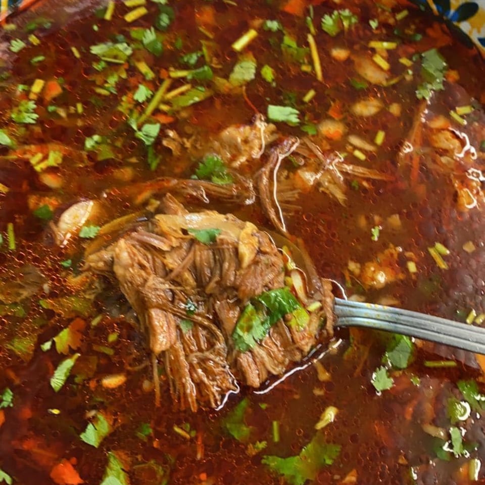 A spoon lifts shredded beef from a rich, dark red Mexican stew with cilantro and spices.