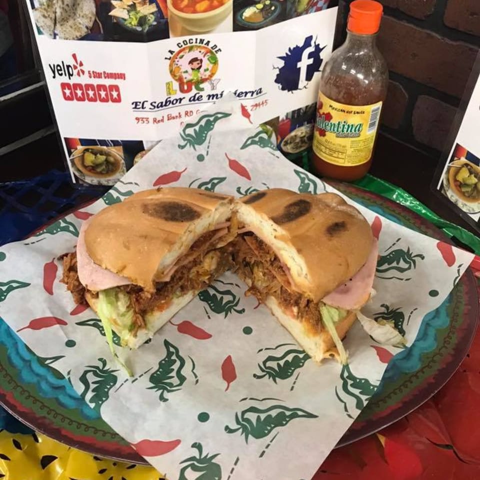 A Mexican torta sandwich, cut in half, revealing shredded meat, ham, and lettuce, with hot sauce and a menu in the background.