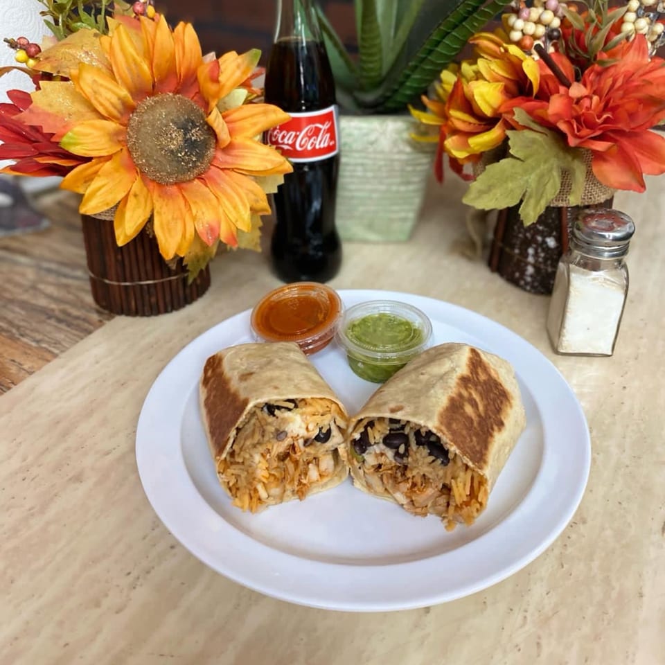 Two burritos cut in half on a white plate with red and green salsa, a Coke, and flowers.