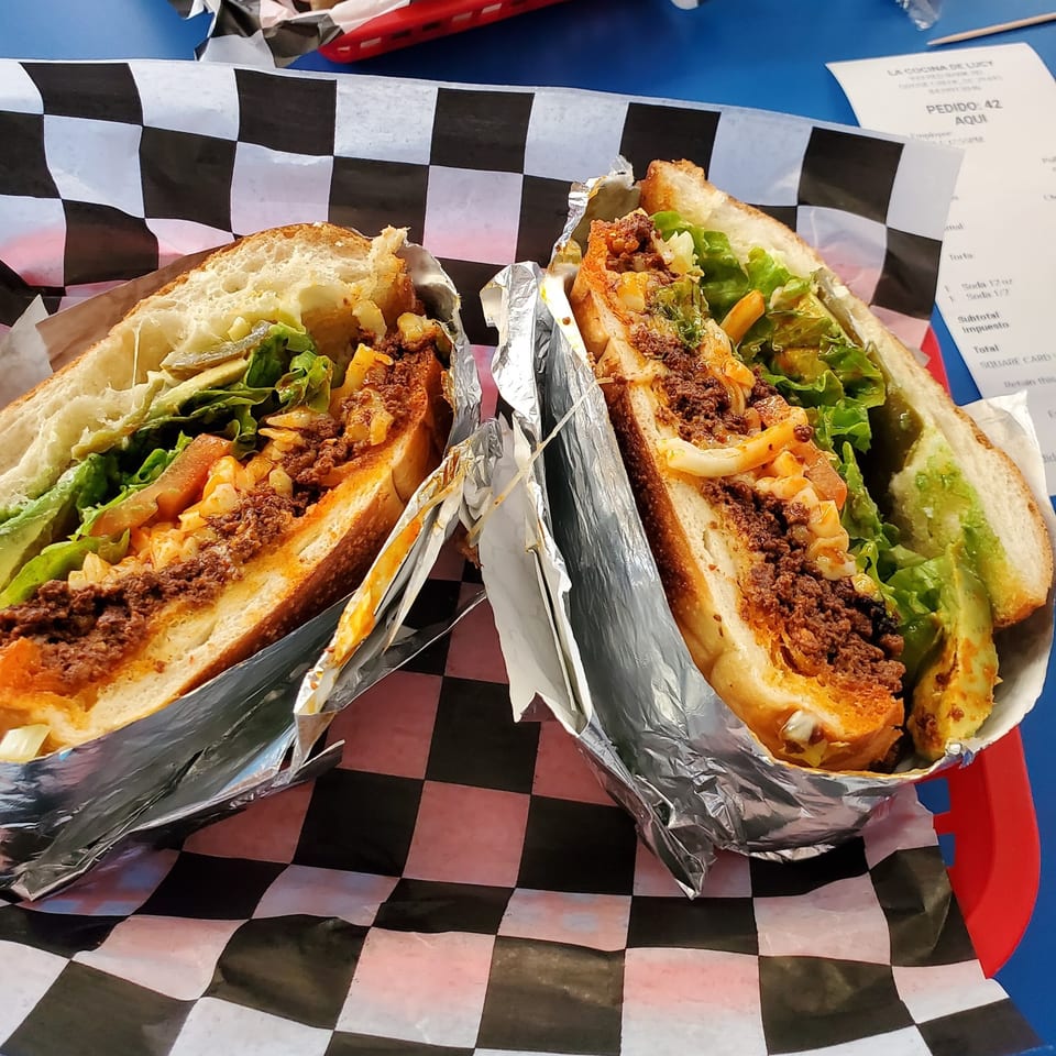Two halves of a Mexican torta sandwich, filled with meat, lettuce, and avocado, on checkered paper.
