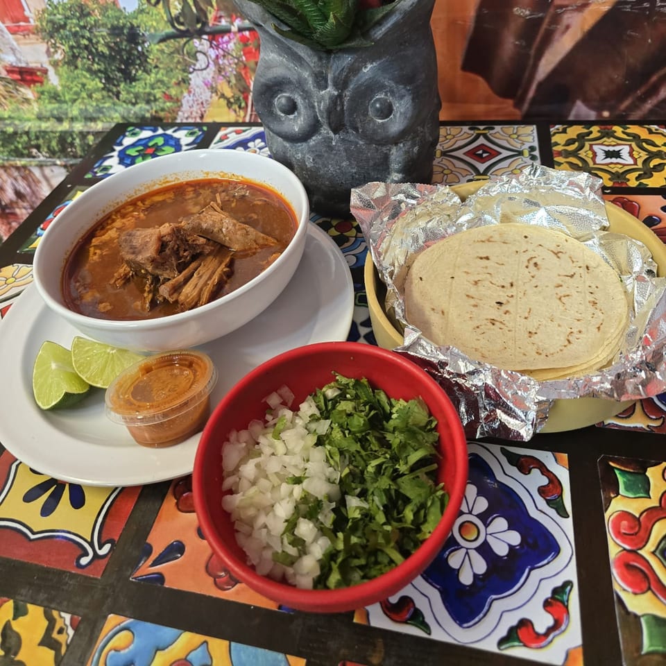 A bowl of birria, limes, salsa, tortillas, and a bowl of chopped onions and cilantro on a colorful tiled table.
