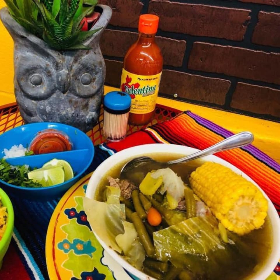 Hearty Mexican soup with corn on the cob, vegetables, and meat, served with lime and cilantro.